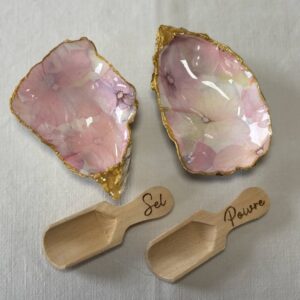 Deux coquilles d’huîtres ornées de motifs d’hortensias roses, dorées et accompagnées de cuillères en bois, dans un coffret ouvert.