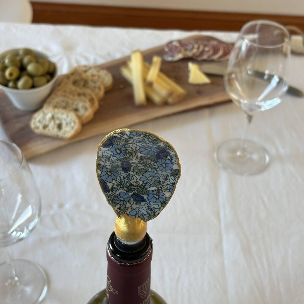 Bouchon de vin original motif petites fleurs bleues, présenté sur une bouteille avec planche apéritive en arrière-plan.